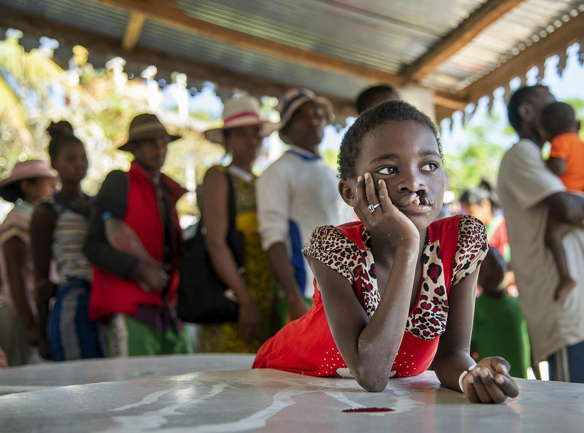 En ung flicka i röd klänning lutar sig mot ett bord med handen mot ansiktet, medan personer står i kö under ett plåttak i bakgrunden.
