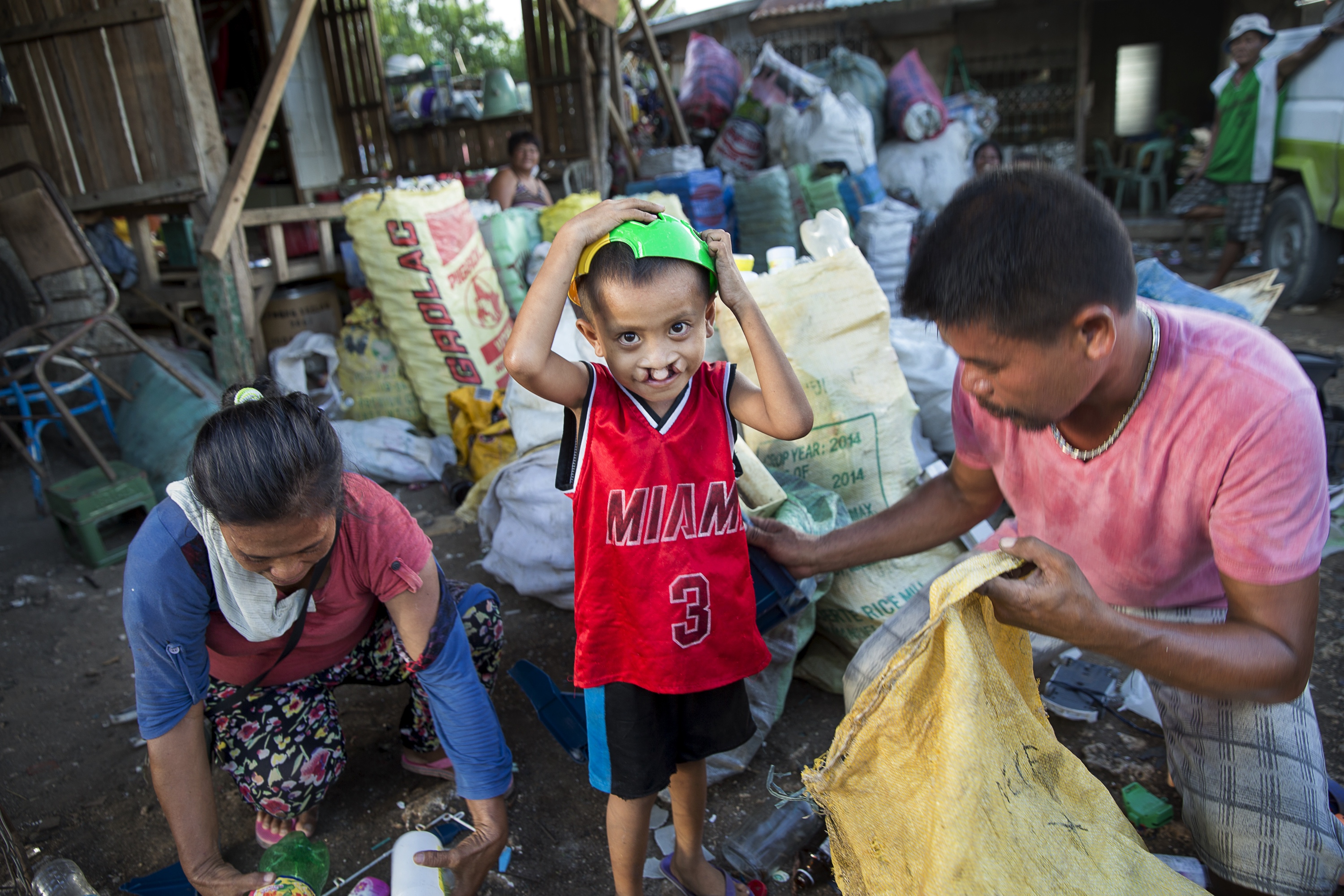 Ett barn med röd t-shirt och grön hink på huvudet, tittar in i kameran med grön hink på huvudet