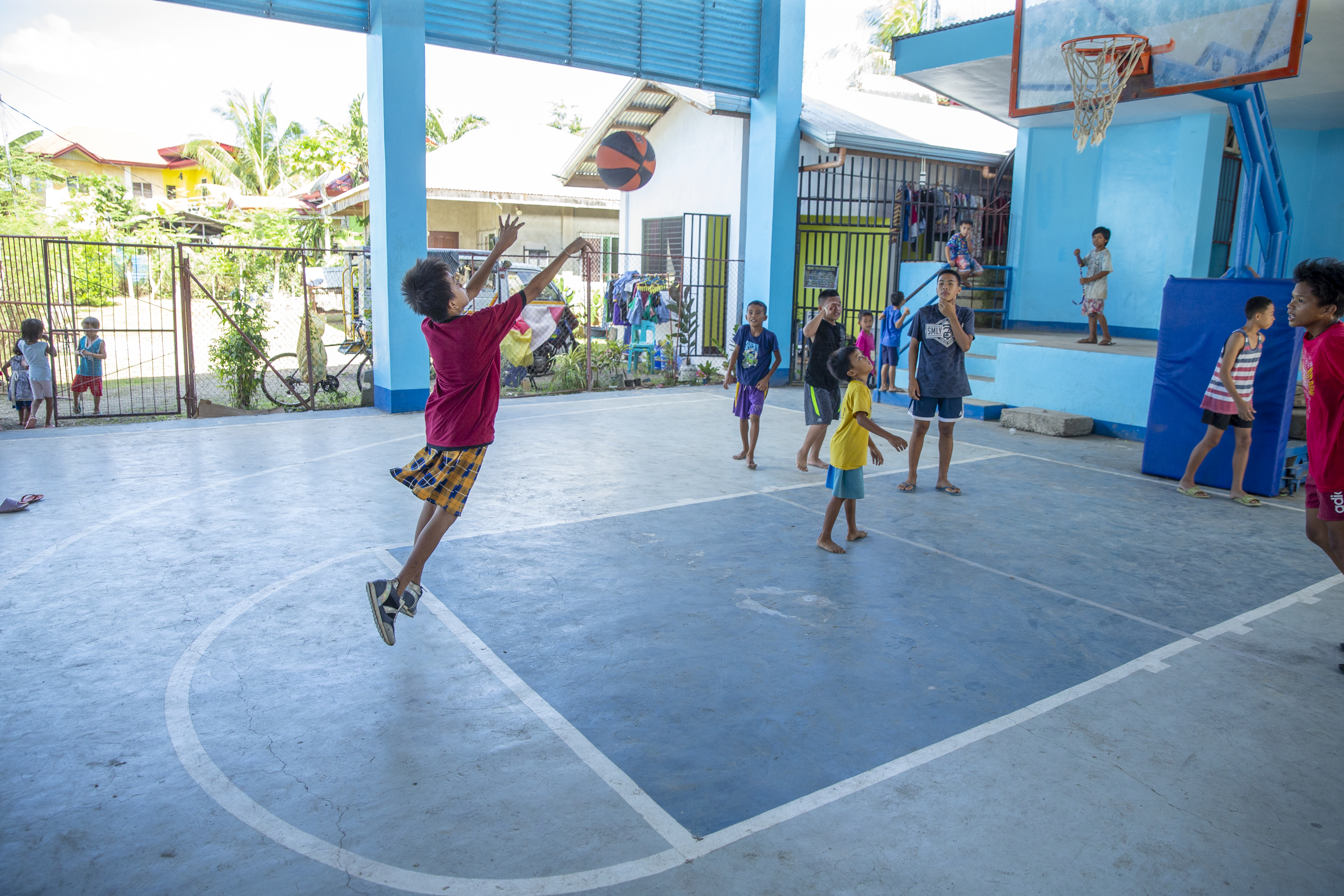 Ett gäng pojkar i olika åldrar spelar basket tillsammans. En av dem hoppar upp i luften och skickar iväg bollen mot korgen.