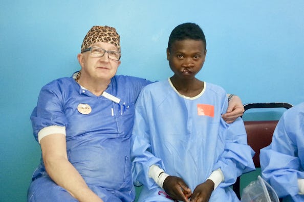 En läkare i sjukhuskläder sitter bredvid en patient i sjukhusrock mot en blå vägg; läkaren har en arm runt patienten.