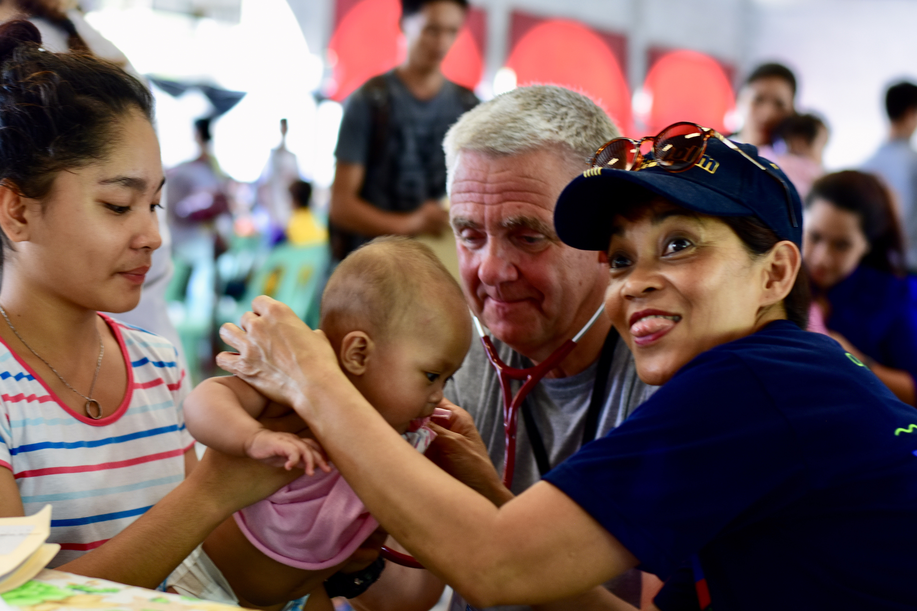 Sjuksköterska i mörkblå t-shirt och keps leker med ett litet barn. Läkare med stetoskop.