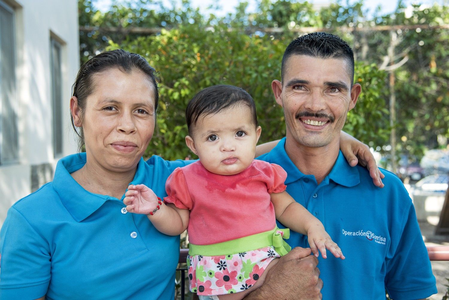 En man och en kvinna båda klädda i blå t-shirt med Operation Smiles logotyp. Mellan sig håller de en liten flicka.