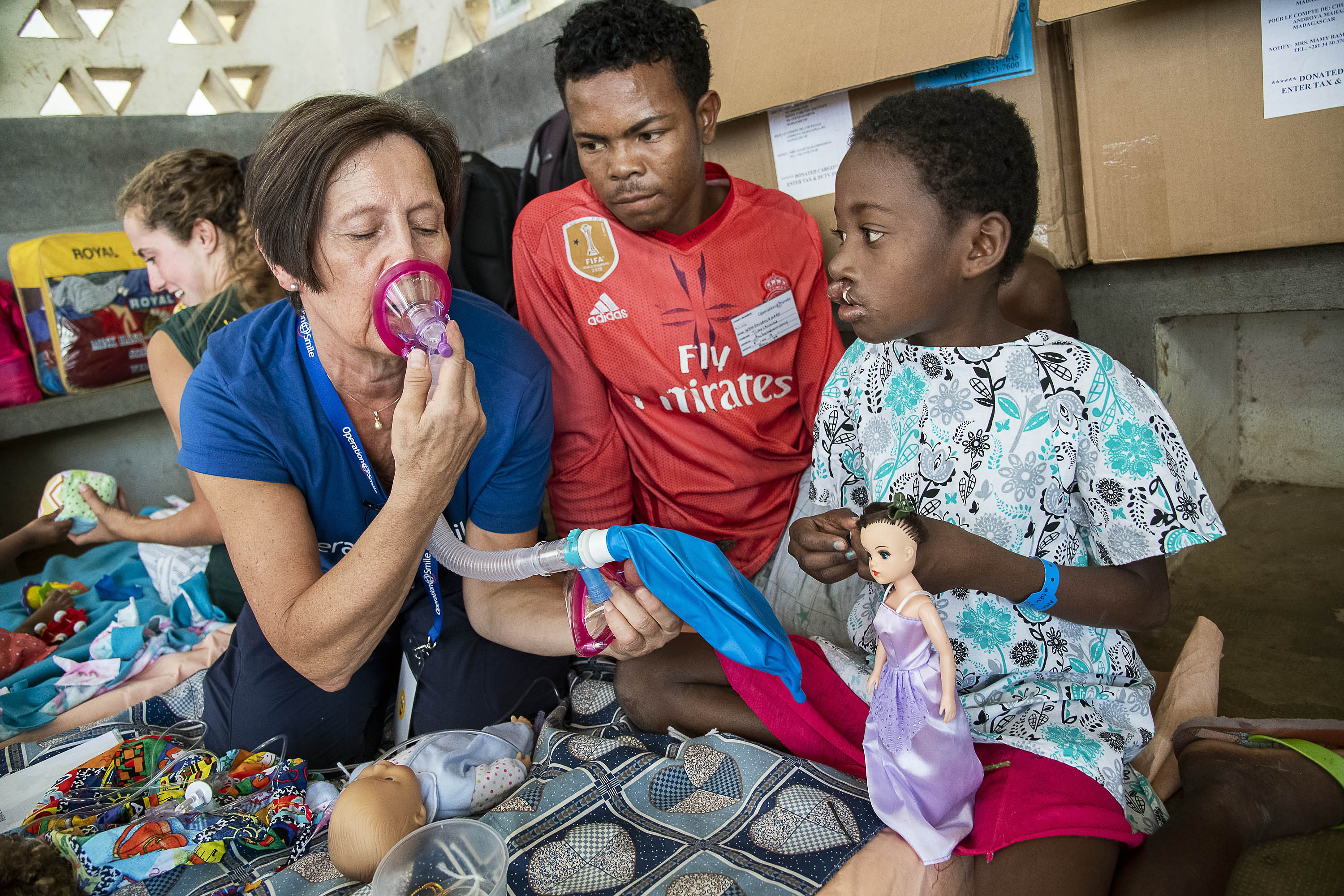 En kvinna visar en medicinsk mask för en pojke och en man. De sitter på golvet med leksaker, dockor, lådor och mattor runt sig.