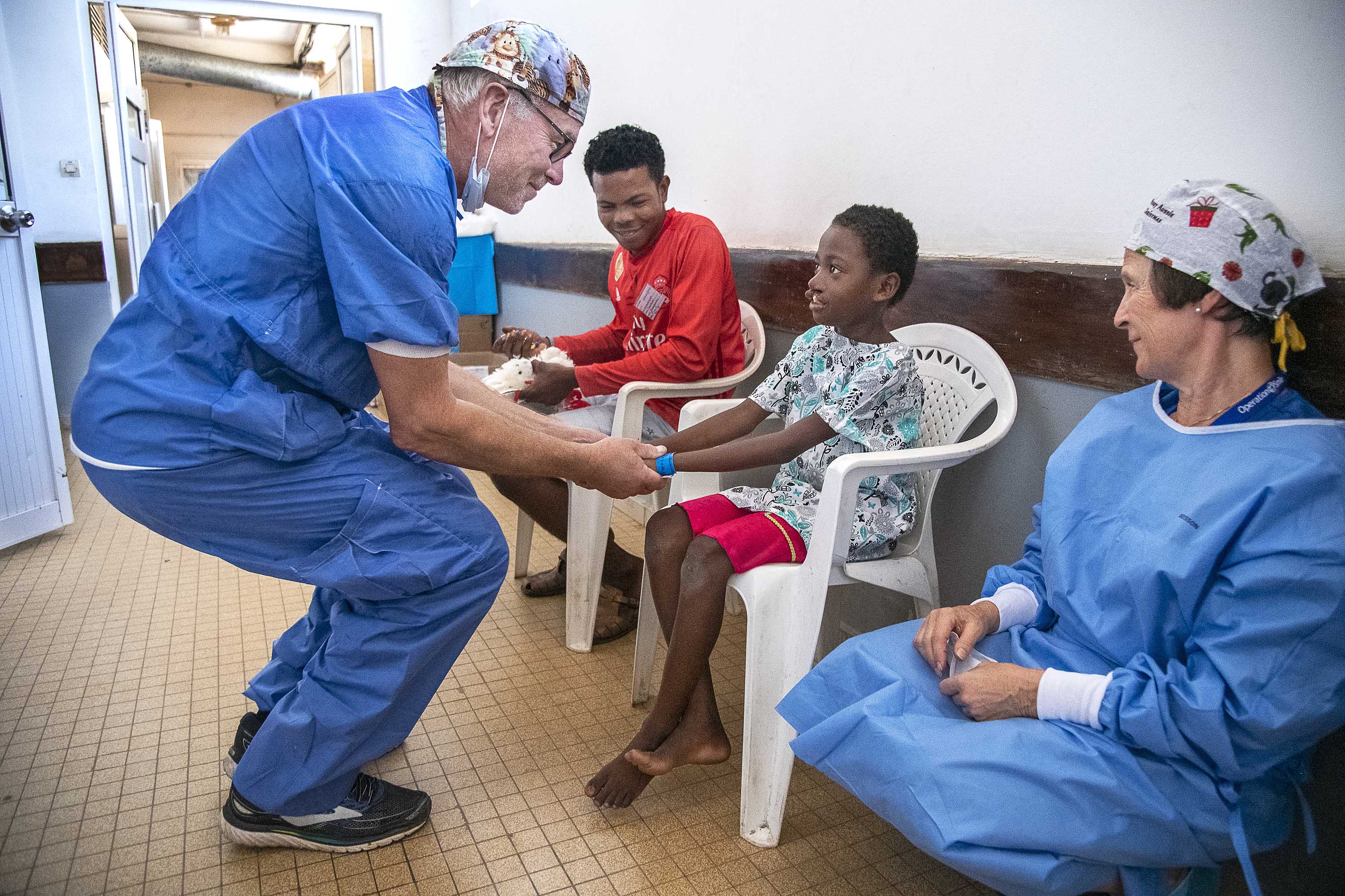 En vårdpersonal i arbetskläder pratar med ett sittande barn, medan ett annat barn och en vuxen tittar på i en klinikkorridor.