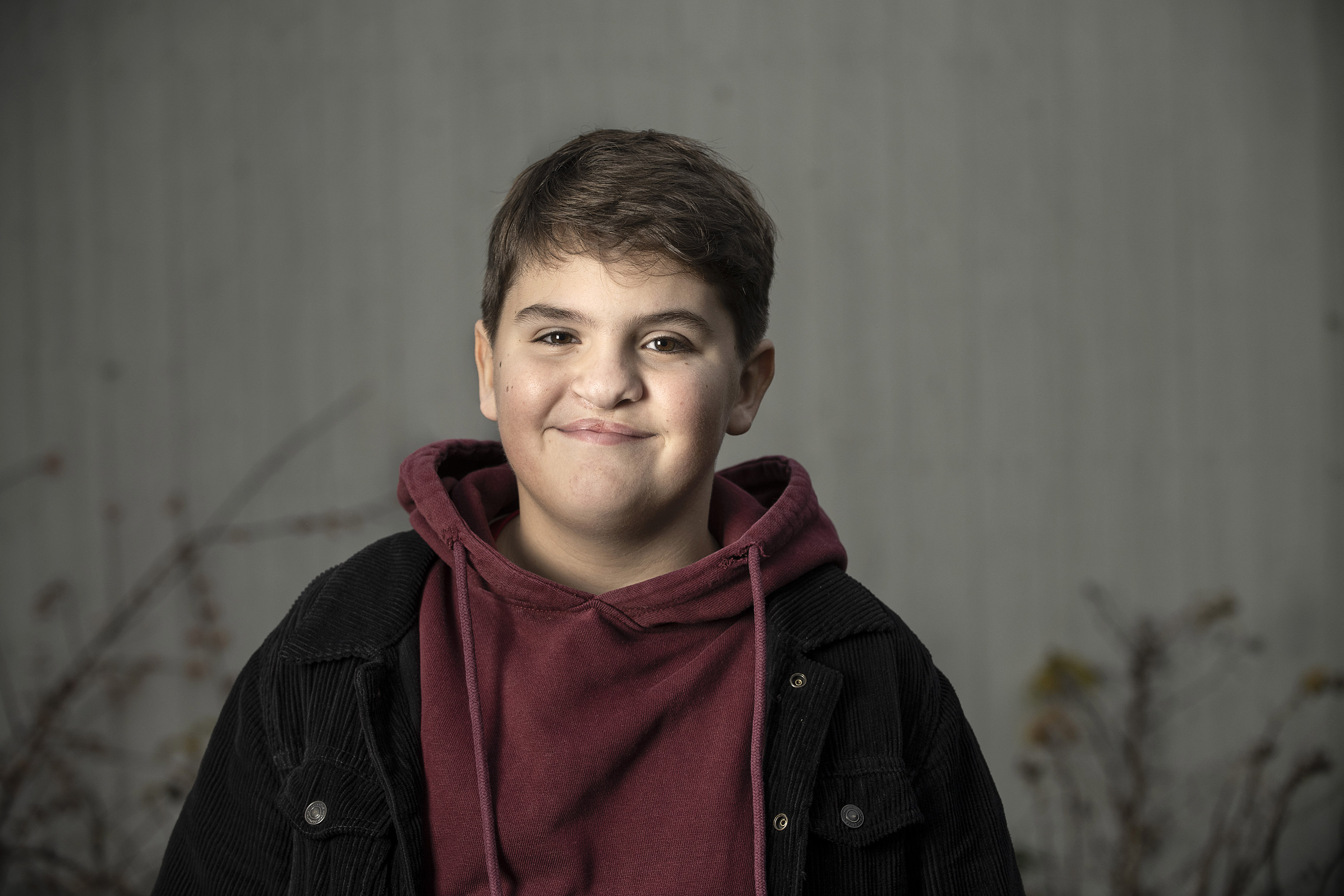 11-årig pojke klädd i vinröd hoodie och svart jacka. Han ser rakt in i kameran och ler med glittrande ögon.