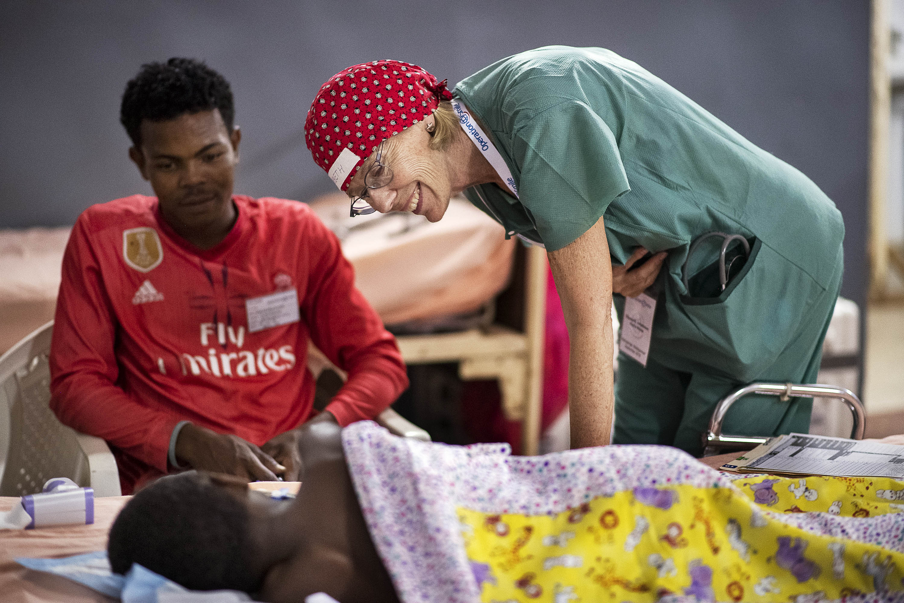 En vårdarbetare i arbetskläder och röd mössa pratar med en patient i sängen, en person i röd tröja sitter bredvid.