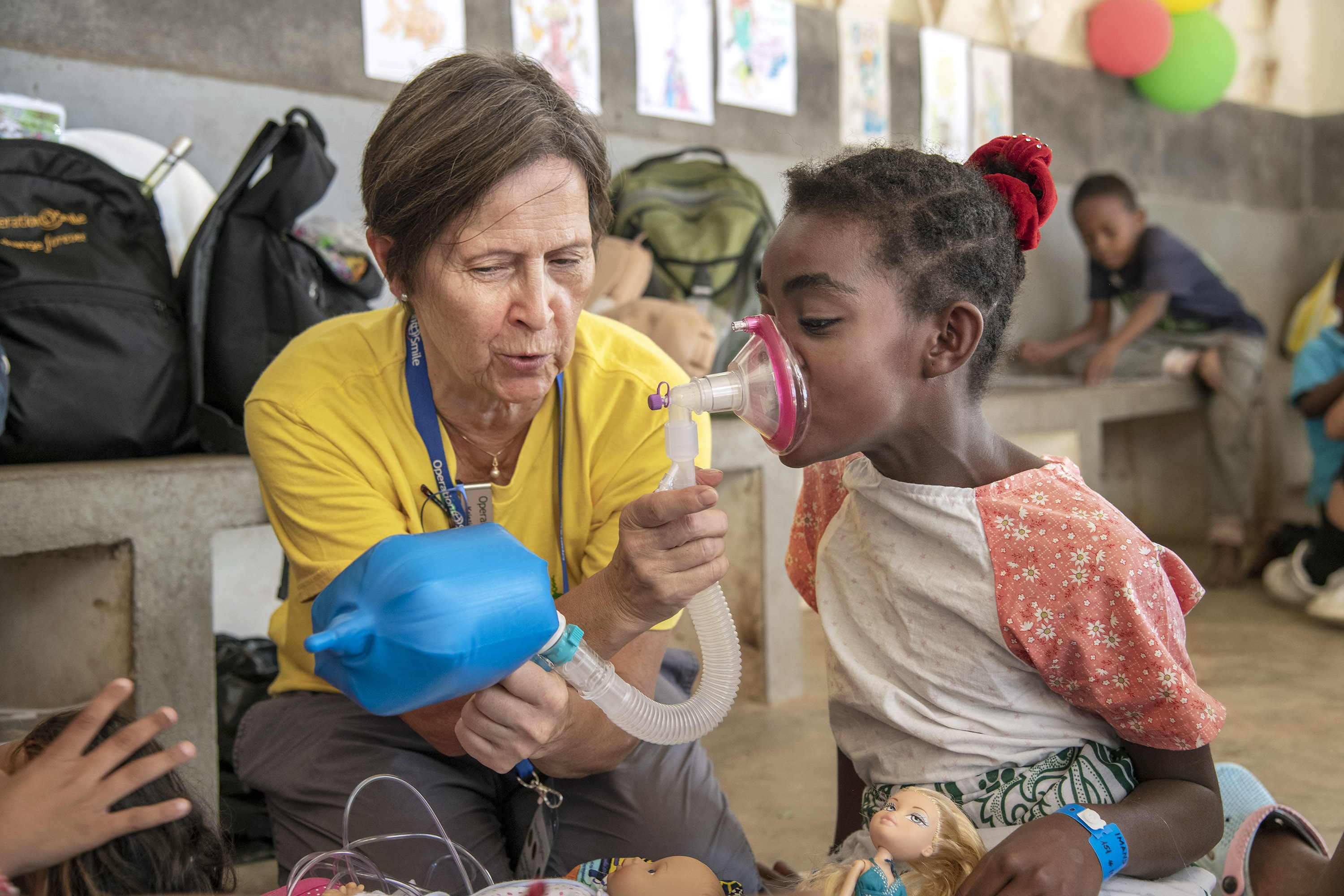 En kvinna i gul t-shirt hjälper till att hålla en narkosmask framför näsa och mun på 10-årig flicka.