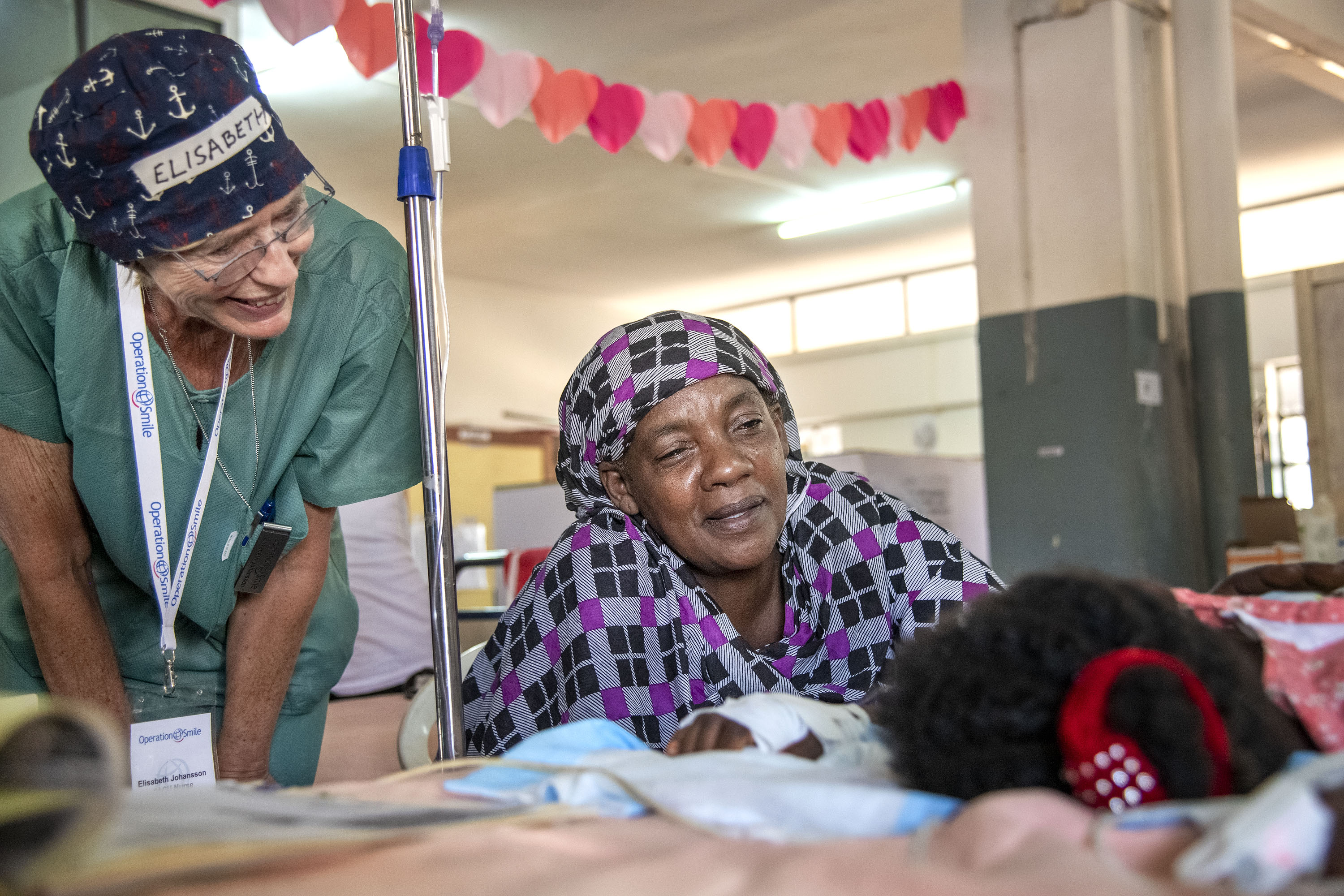 En äldre kvinna sitter bredvid en sjukhussäng där man skymtar huvudet på patienten. Bredvid kvinnan står en kvinna iklädd gröna sjukhuskläder. I bakgrunden, i taket hänger en girland med hjärtan.