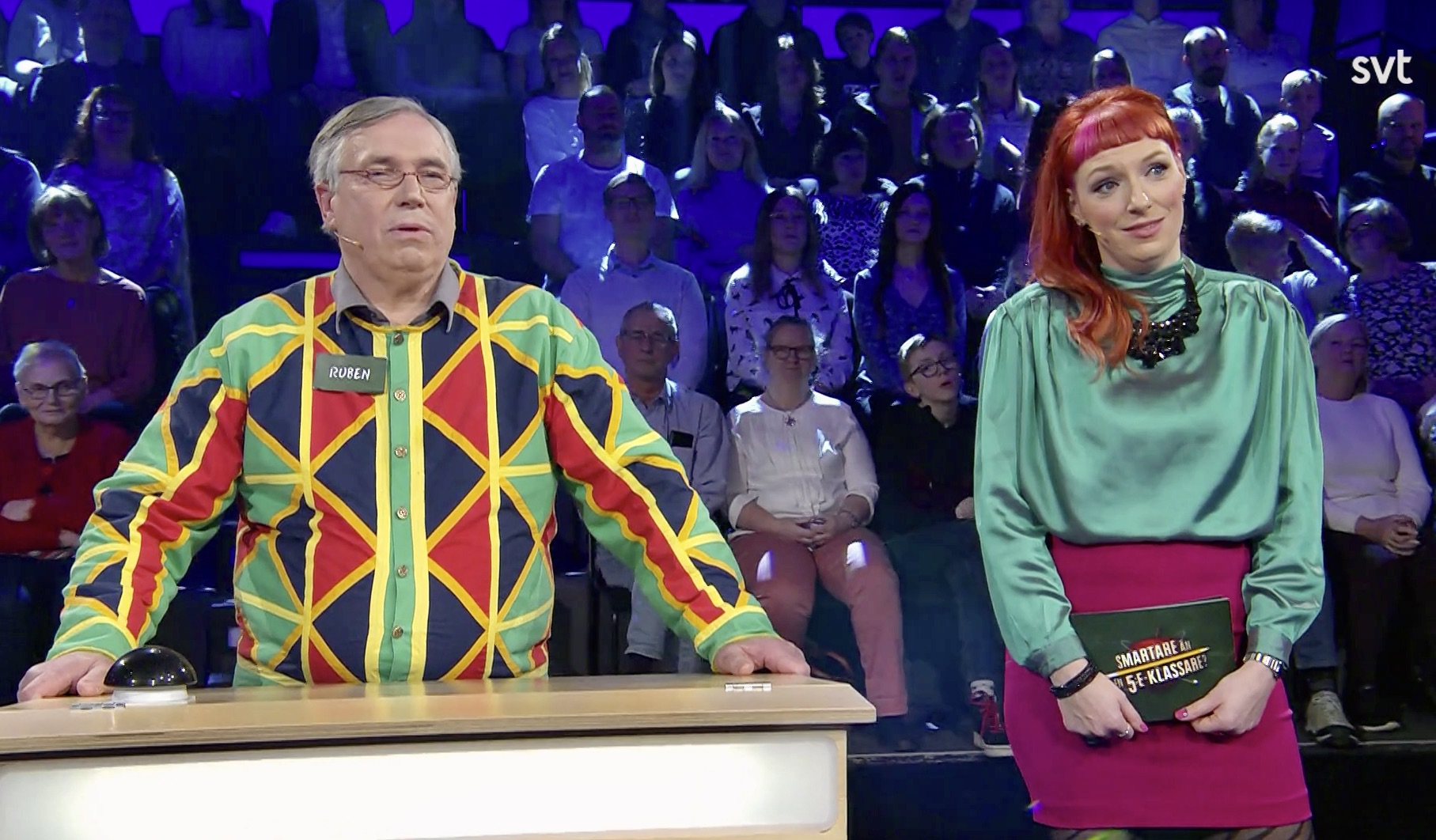 Bild från tv där en man i en stormönstrad skjorta står bredvid programledaren. De båda tittar åt samma håll, förbi kameran.
