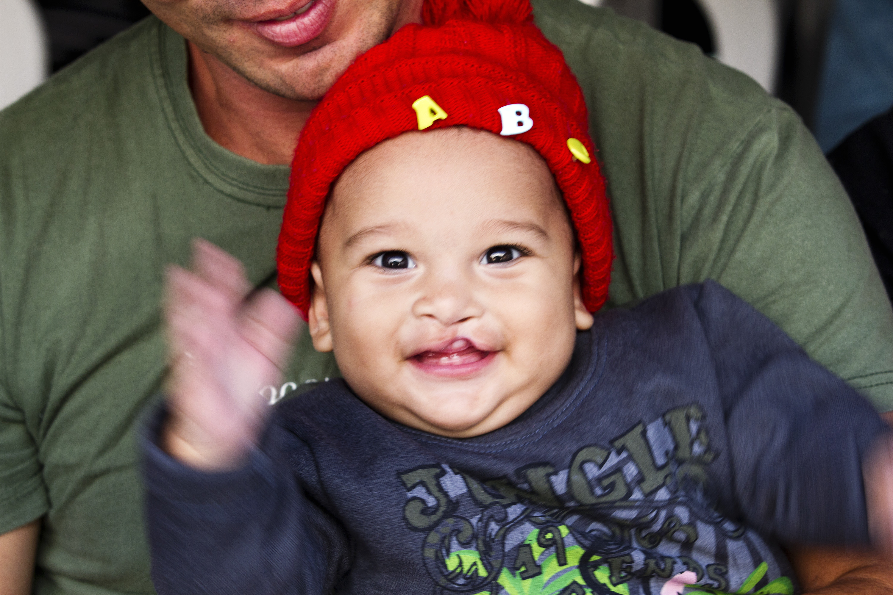 En bebis med röd stickad mössa med bokstäver sitter i knät på en vuxen och vinkar med händerna.