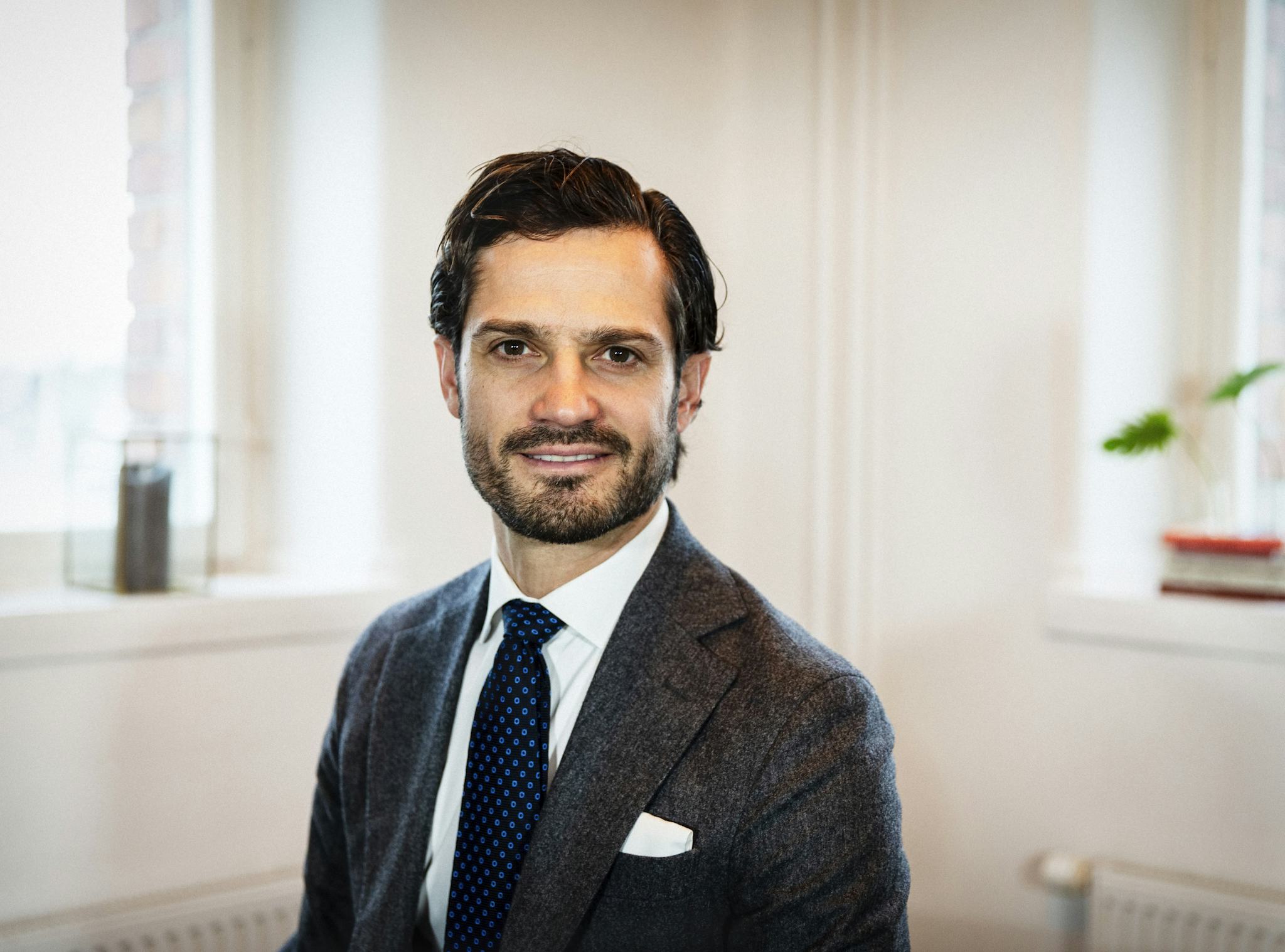 Prins Carl-Philip. En man med mörkt hår och skägg, iklädd kostym och slipsar, sitter inomhus framför två fönster med vita väggar och några saker på fönsterbrädorna.