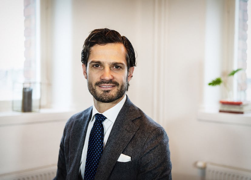 Prins Carl-Philip. En man med mörkt hår och skägg, iklädd kostym och slipsar, sitter inomhus framför två fönster med vita väggar och några saker på fönsterbrädorna.