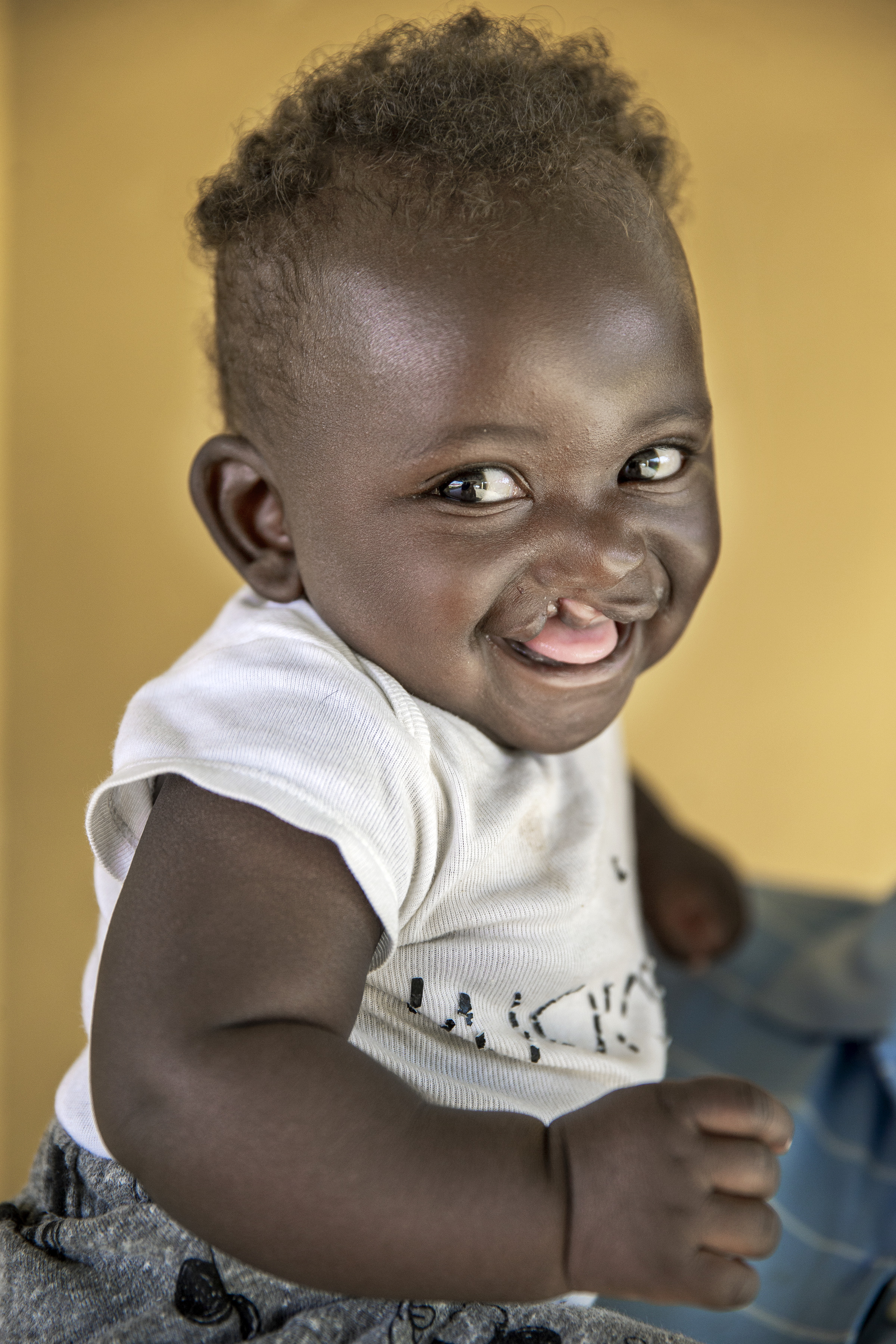 Ett barn med lockigt hår ler och räcker ut tungan, har på sig en vit tröja och sitter inomhus mot en gul bakgrund.