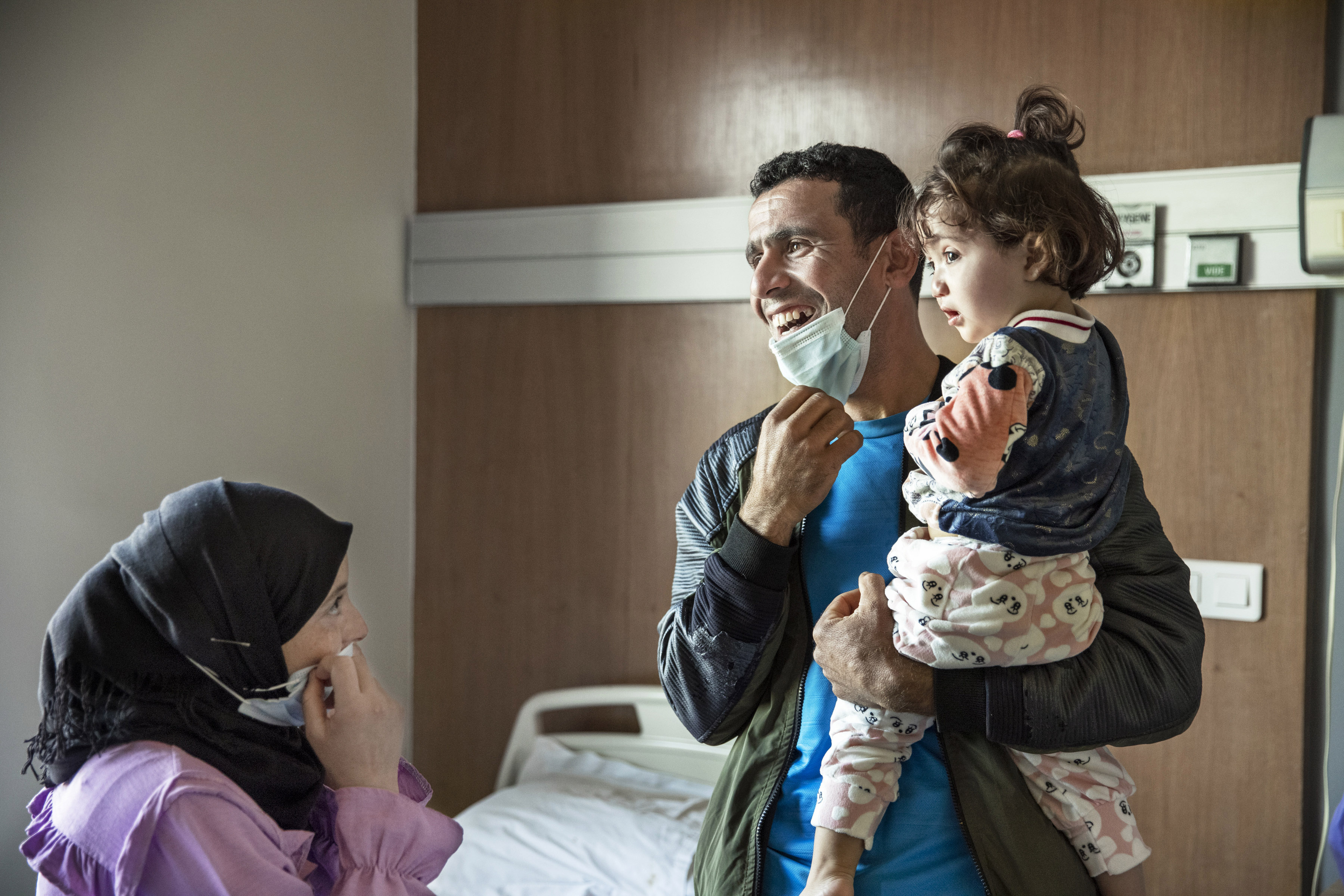En man håller sin dotter, ca 2 år gammal, i famnen. De befinner sig i en sjukhussal. I det vänstra nedre hörnet syns en kvinna, flickans mamma.