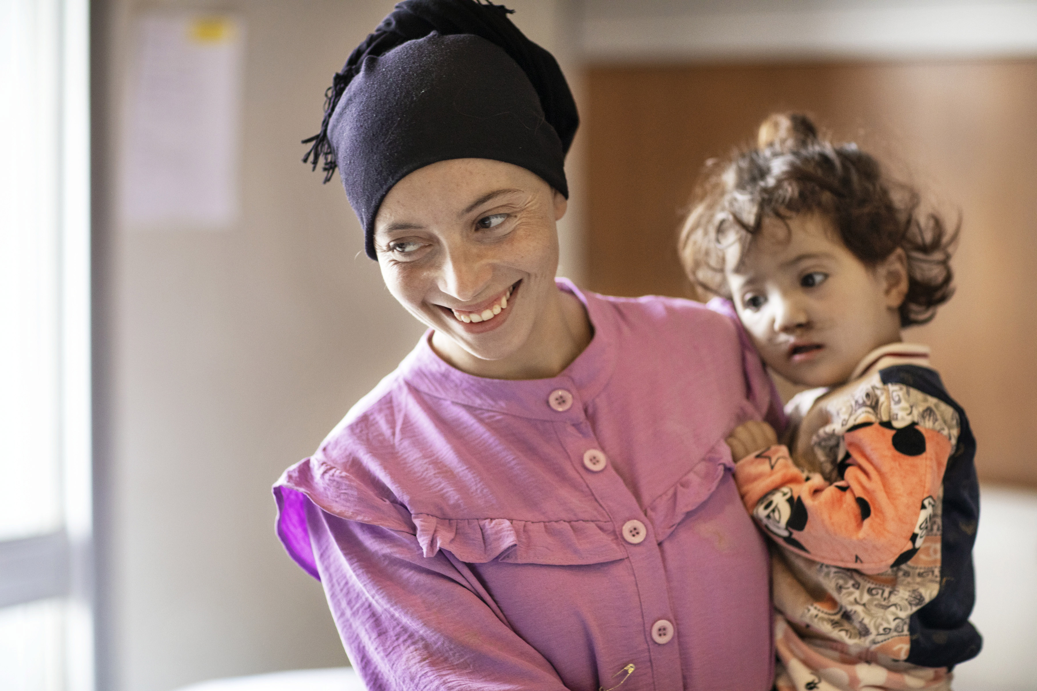 En kvinna i rosa blus och med en svart sjal om håret håller sin dotter i famnen. De tittar båda på något utanför bilden och kvinnan ler.