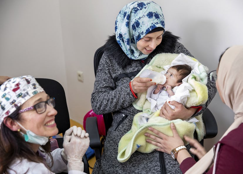 En kvinna håller sin baby i famnen och matar henne med hjälp av en nappflaska. PÅ golvet framför sitter ytterligare två kvinnor, varav en är tandläkare.