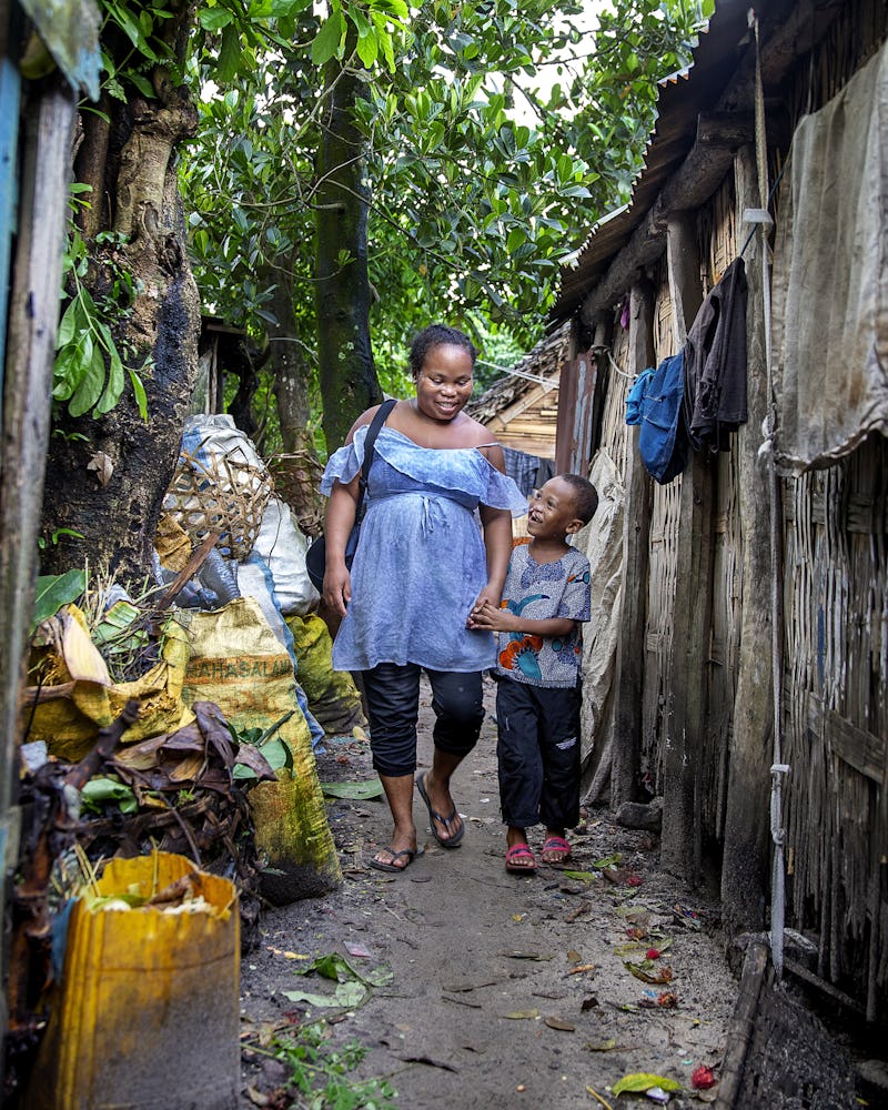 En kvinna och ett barn går genom en smal gränd mellan enkla träbyggnader, omgivna av hushållsföremål och växter.