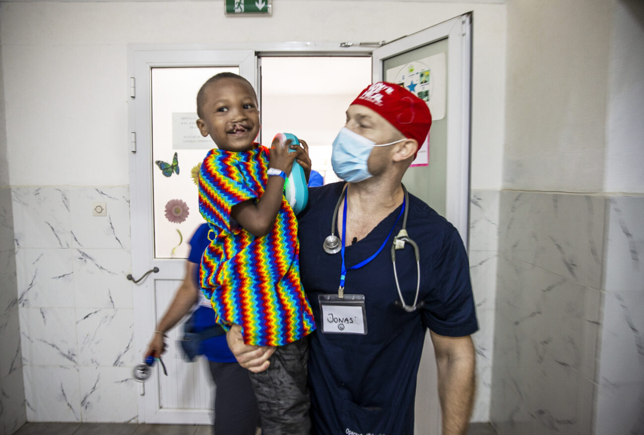 Bild på en glad sexårig Honoré som har en leksak i handen. Han är iklädd en färgglad randig operationsskjorta i armarna på narkosläkare Jonas Wanbro som är iklädd operationskläder och munskydd.
