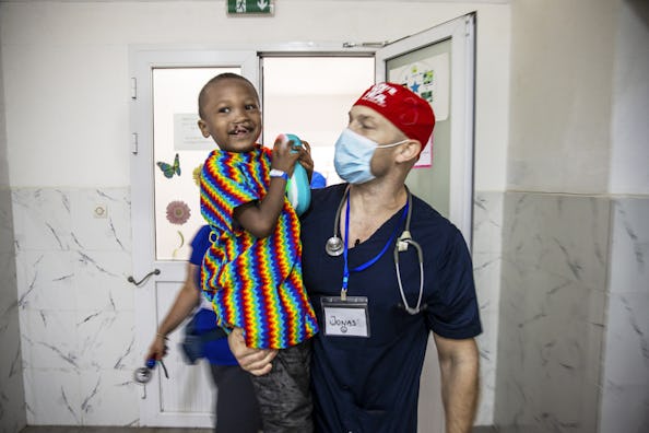 En glad sexårig Honoré som har en leksak i handen. Han är iklädd en färgglad randig operationsskjorta i armarna på narkosläkare Jonas Wanbro som är iklädd operationskläder och munskydd.