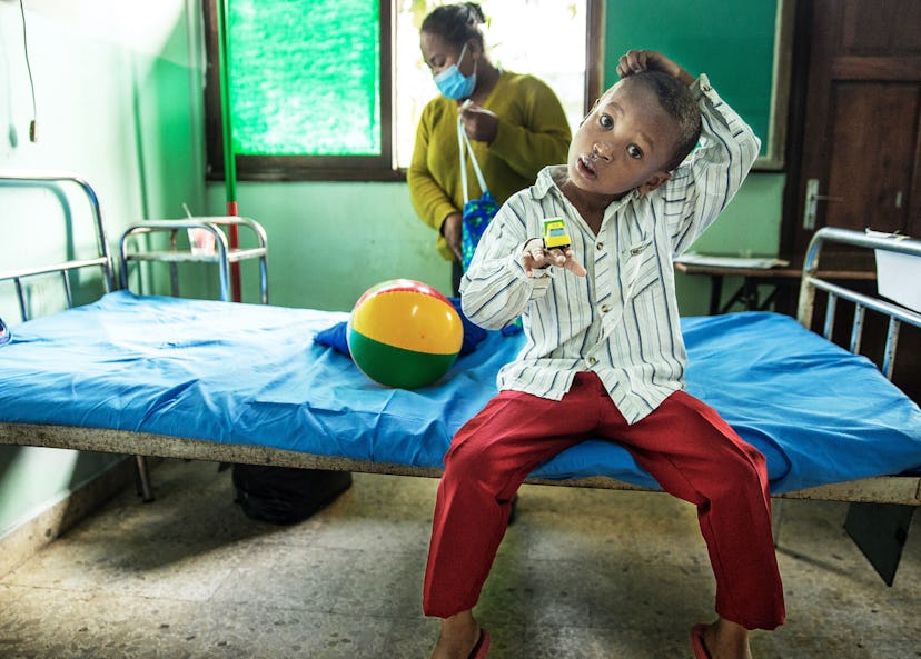En sexårig pojke sitter på en sjukhussäng med huvudet lite på sned. Han balanserar en leksaksbil på sin hand och har en boll bredvid sig. I bakgrunden syns en kvinna som plockar ned saker i en väska.