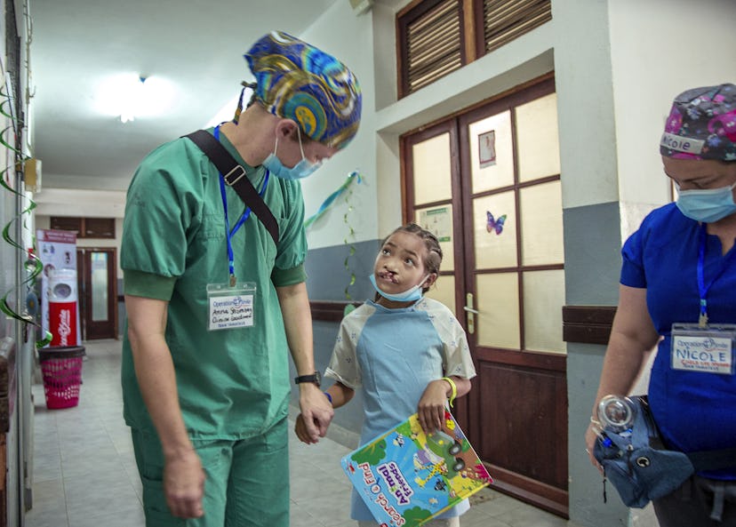 En vårdpersonal i arbetskläder håller handen på en ung patient med en bok, medan en annan personal går förbi i sjukhuskorridoren.