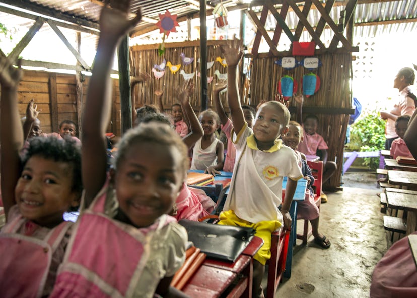 Honoré sitter i sin skolbänk i klassrummet och räcker upp handen. Lokalen är spartanskt inredd och har barnens teckningar på väggen.