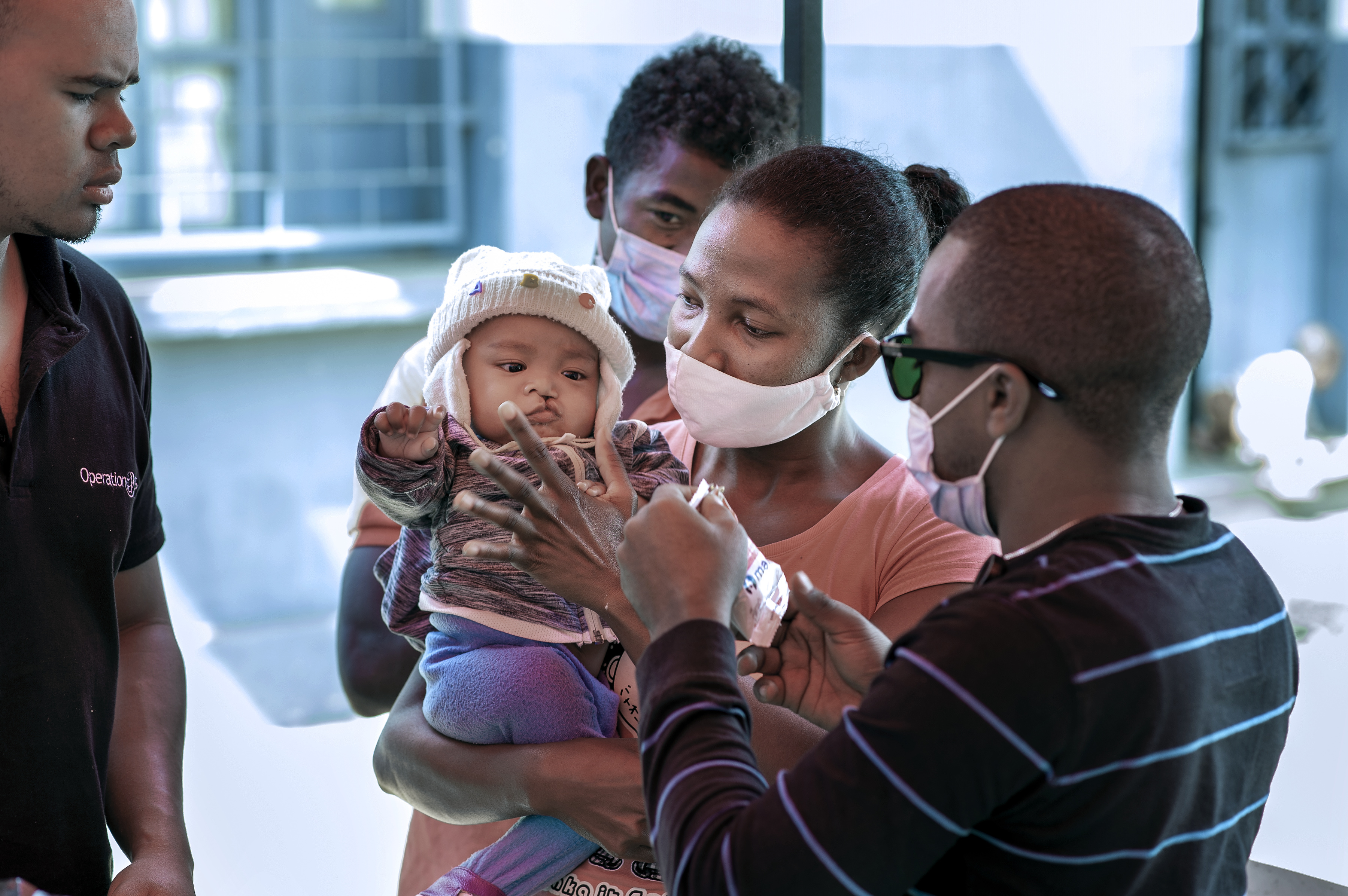 En kvinna med munskydd håller i en bebis medan en man ger vaccin; två andra vuxna ser på inomhus.