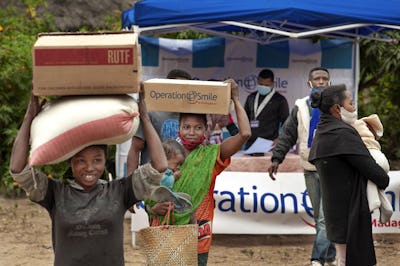 Personer bär lådor och förnödenheter framför ett Operation Smile-tält; några har munskydd och håller barn, vid en utdelning.