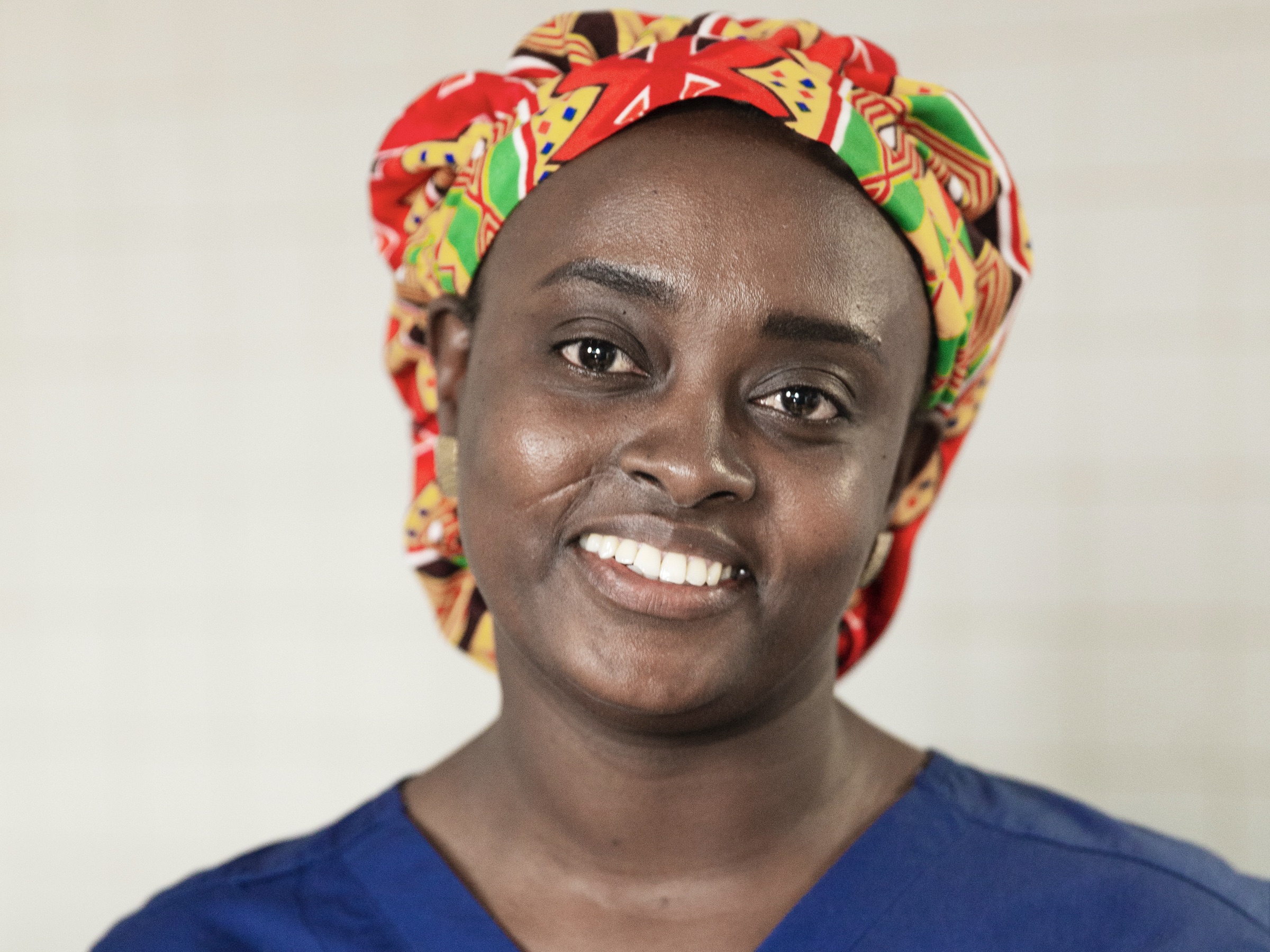 En kvinna med mönstrad huvudduk och blå tröja ler mot kameran i en inomhusmiljö med bra belysning.