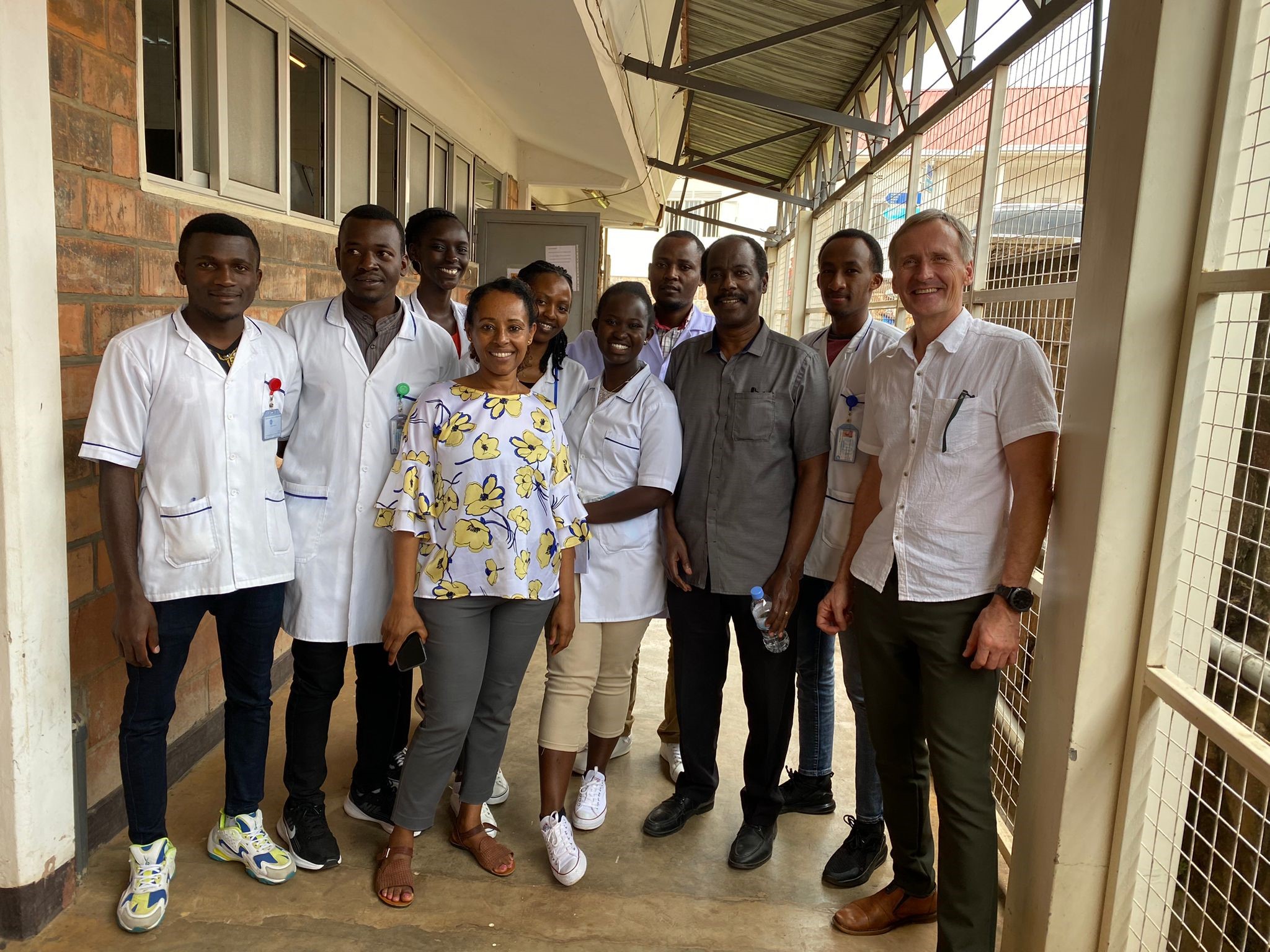 En grupp på tio personer, några i vita läkarrockar, står tillsammans och ler mot kameran i en korridor med metallräcken och tak.