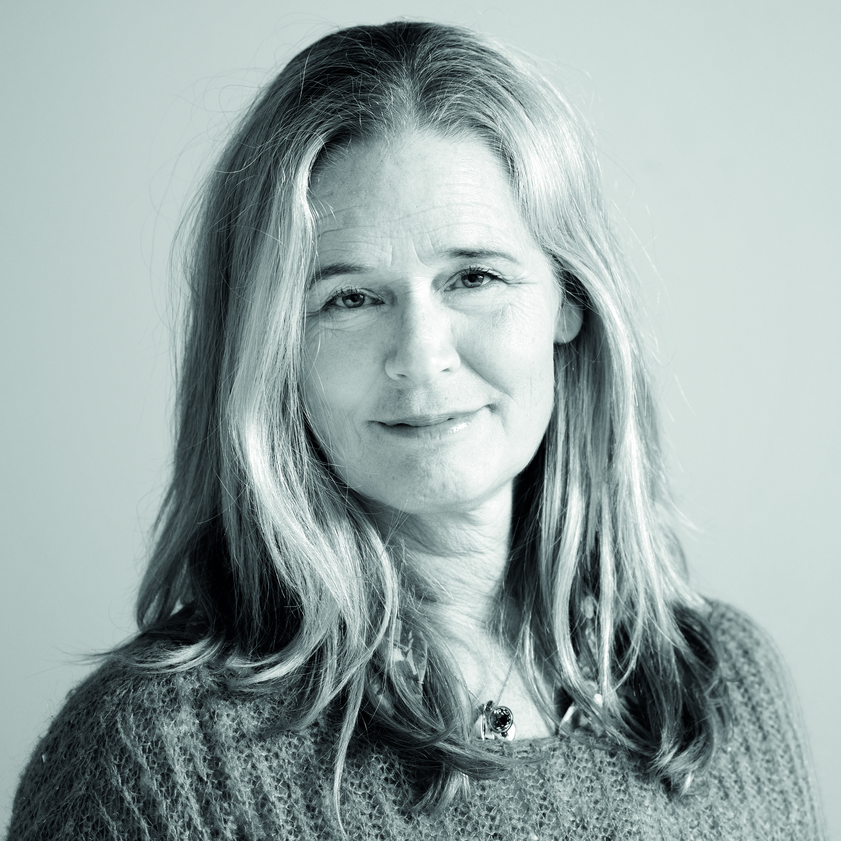 En kvinna med långt, ljust hår och en tröja tittar mot kameran med neutral min framför en enkel bakgrund.