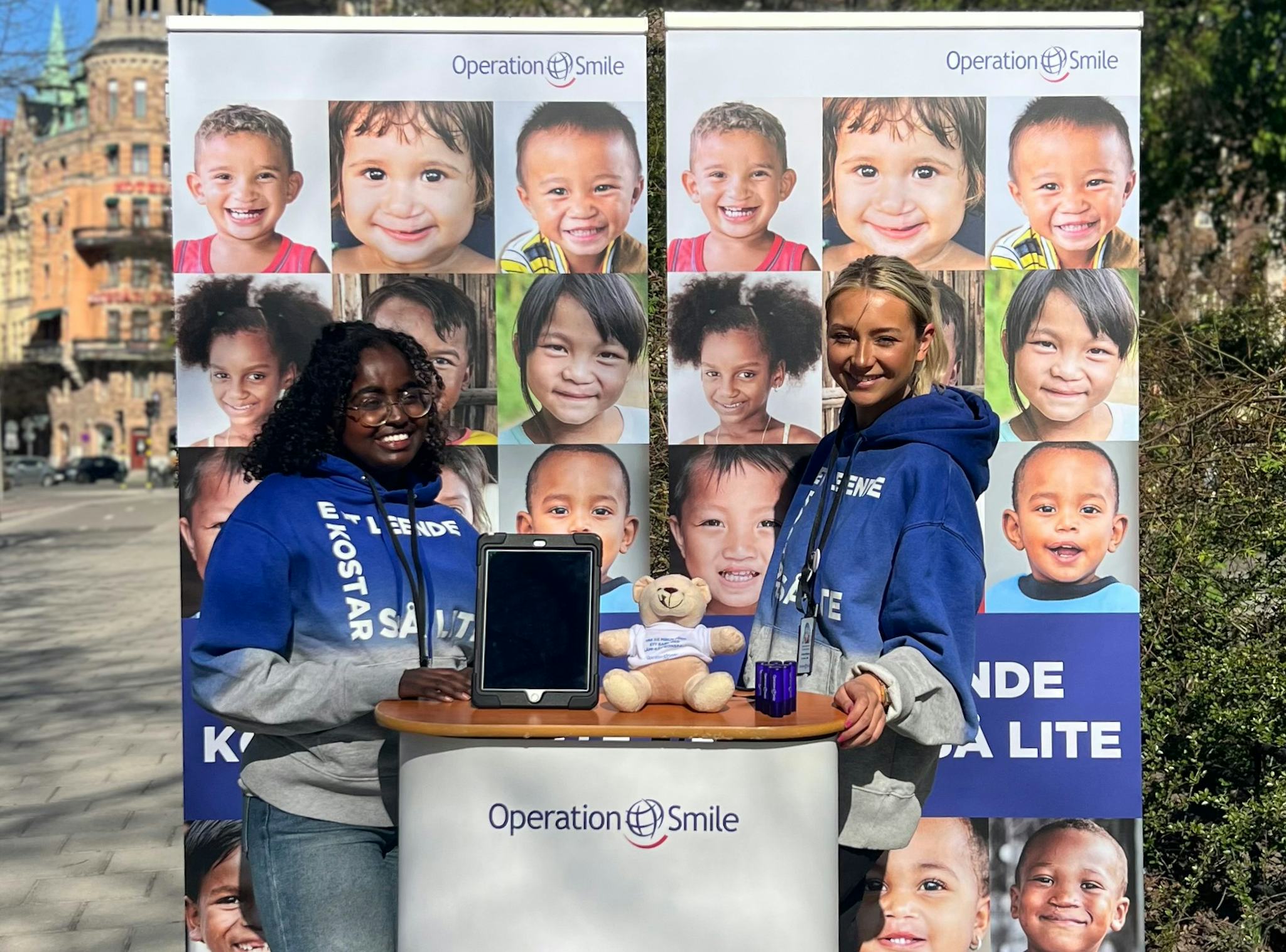 Två kvinnor står vid ett Operation Smile-bås utomhus med bilder på leende barn i bakgrunden och en nalle, broschyrer samt en surfplatta på bordet.