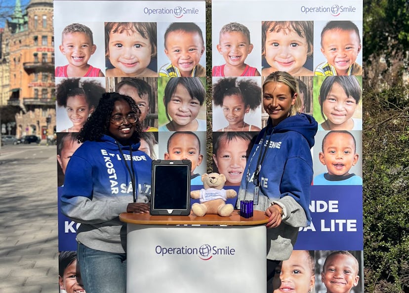 Två kvinnor står vid ett Operation Smile-bås utomhus med bilder på leende barn i bakgrunden och en nalle, broschyrer samt en surfplatta på bordet.