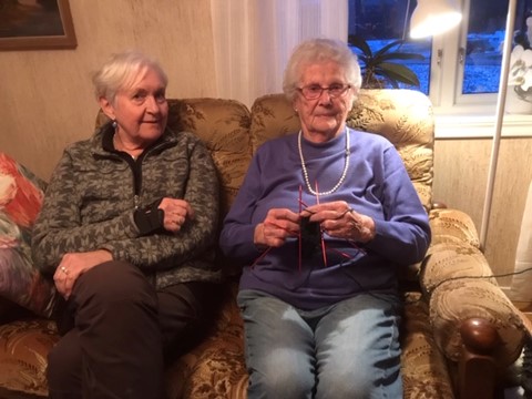 Två kvinnor sitter bredvid varandra i en soffa. Den ena stickar, den andra tittar på.