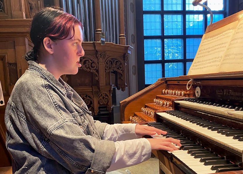 En person i jeansjacka spelar ett orgel och läser noter i ett rum med trädetaljer och stora fönster.