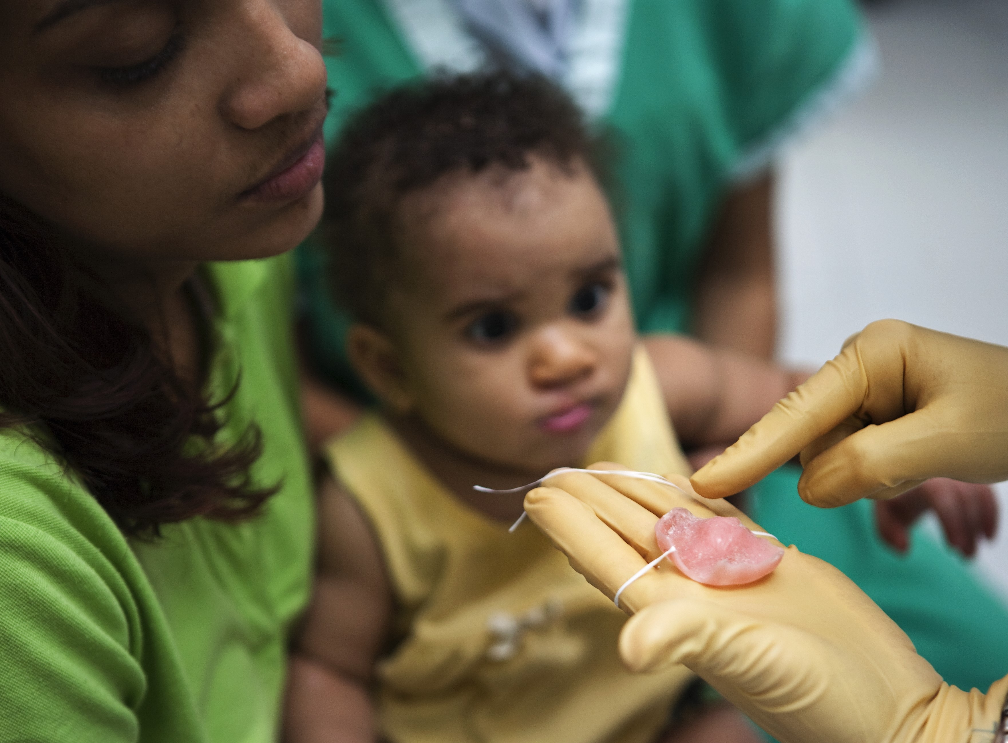 En gomplatta ligger i en hand med plasthandske framför en liten patient som sitter i sin mammas knä. Foto: Marc Ascher