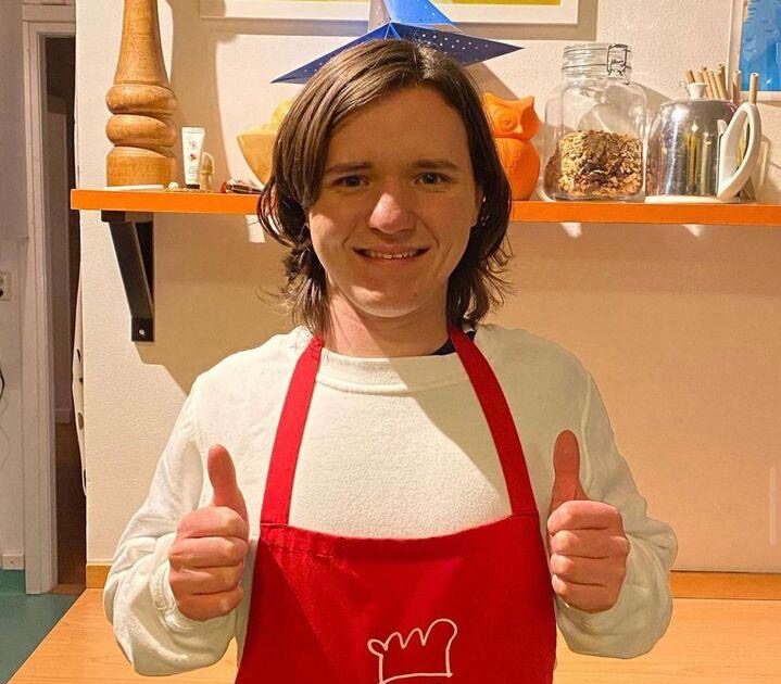 En person med röd förkläde och vit skjorta står inomhus och visar två tummar upp framför en kökshylla.