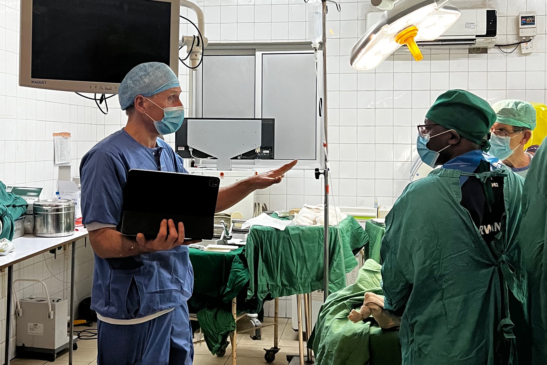 En kirurg med en bärbar dator pratar med två vårdpersonal i en operationssal, alla har operationskläder, masker och mössor.