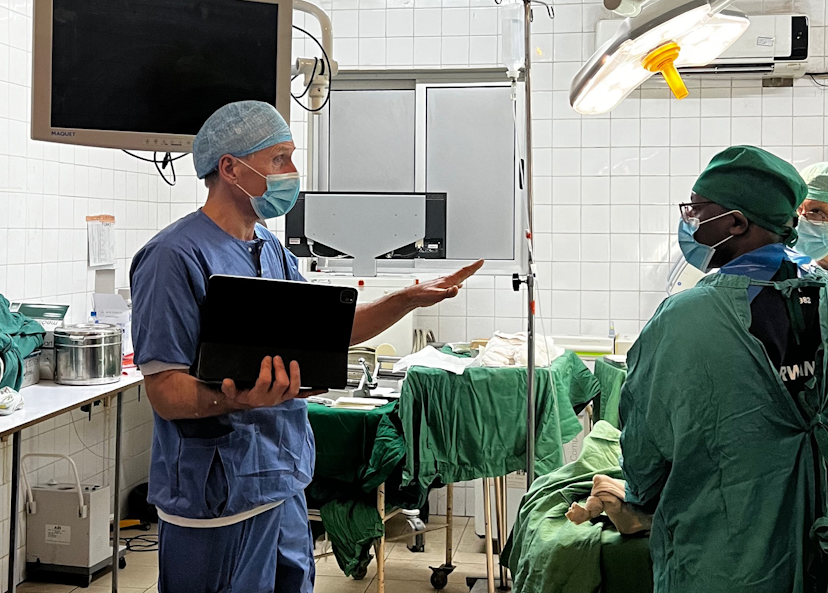 En kirurg med en bärbar dator pratar med två vårdpersonal i en operationssal, alla har operationskläder, masker och mössor.