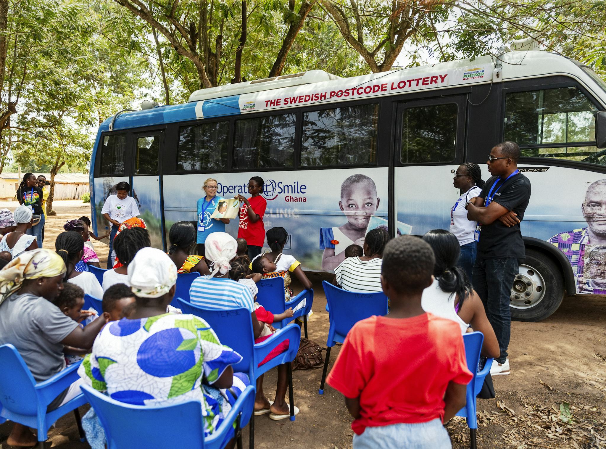 En grupp människor sitter på plaststolar utomhus under några träd. De lyssnar till 2 personer som föreläser. I bakgrunden syns en buss .