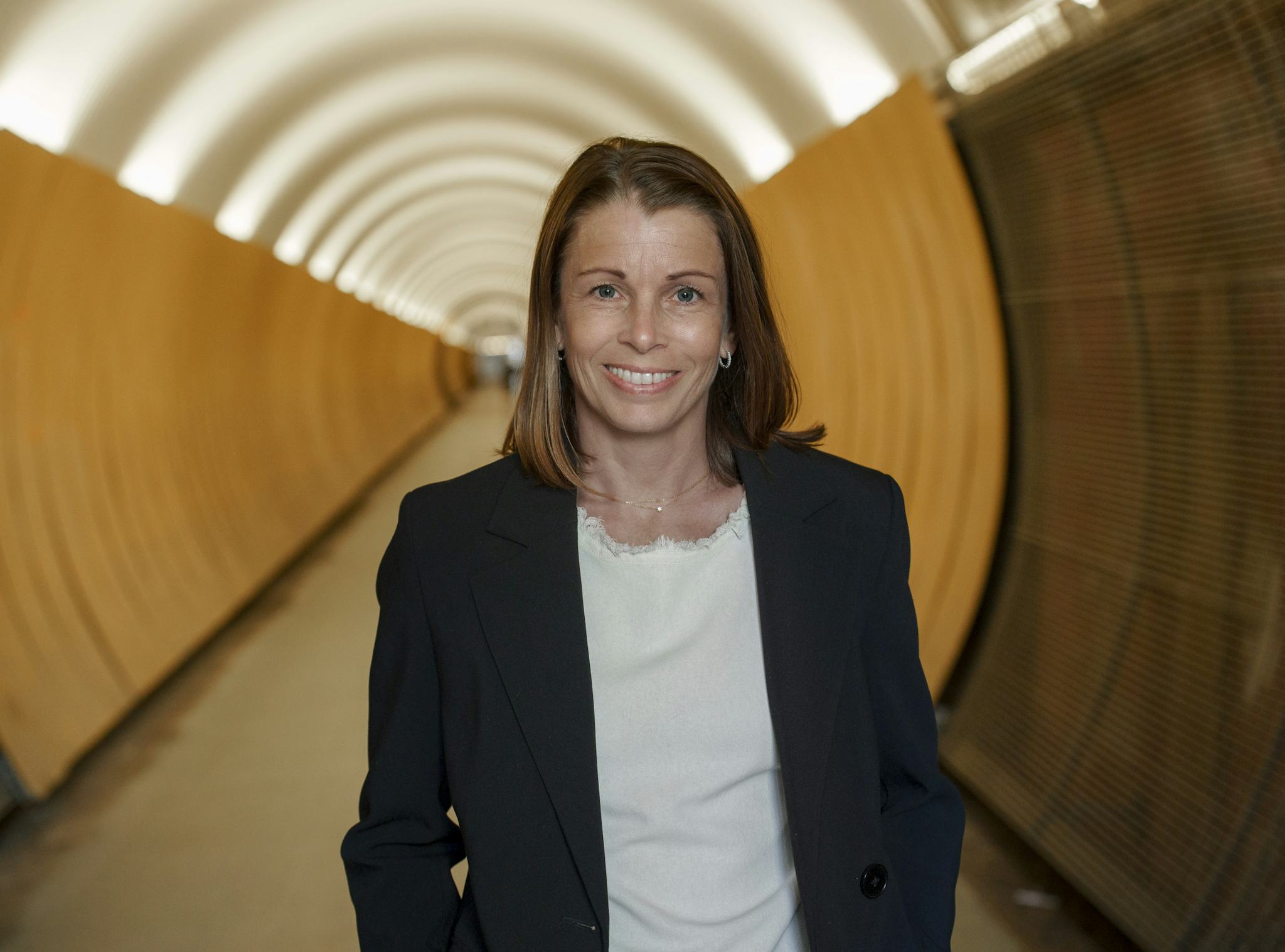 En leende Malin Söderström med axellångt brunt hår och klädd i mörk kavaj och vit blus. Hon står framför en lång gul gångtunnel i vilken ljuset skapar ett intressant mönster i taket.