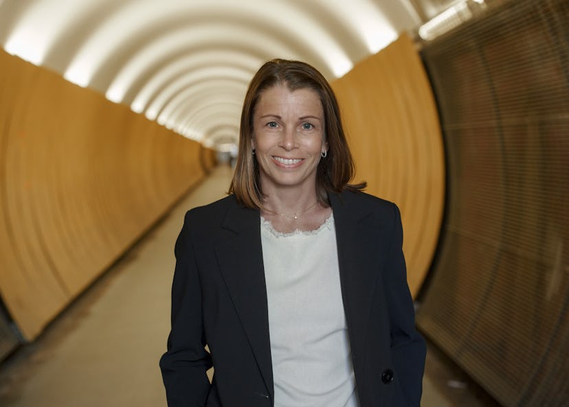 En leende Malin Söderström med axellångt brunt hår och klädd i mörk kavaj och vit blus. Hon står framför en lång gul gångtunnel i vilken ljuset skapar ett intressant mönster i taket.