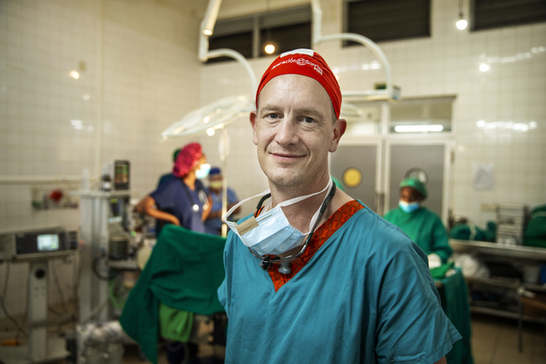 En man tittar in i kameran. Han är iklädd operationskläder och en röd operationsmössa. Han har ett munskydd och pannlampa hängade runt halsen. Bakom skymtar ett operationsbord och sjukvårdspersonal.
