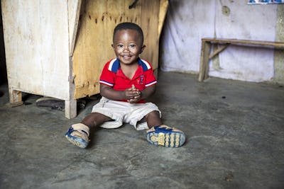 En glad pojke i tvåårsåldern sitter på golvet. Han har en röd t-shirt med blåvit krage, krämfärgade korta byxor och sandaler.