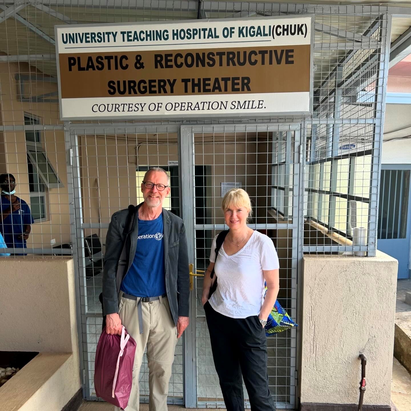 En man och en kvinna, båda volontärer för Operation Smile, står framför ingången till en byggnad. På skylten bakom dem står det University Teaching Hospital of Kigali, Plastic & Reconstructive Surgery Theater, Courtesy of Operation Smile.