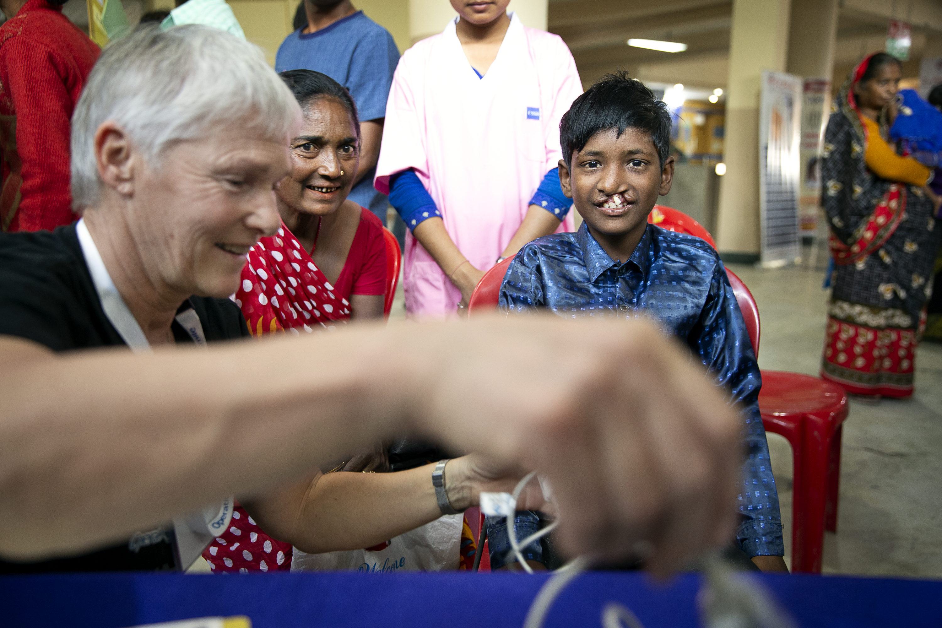 Tirth, en pojke i yngre tonåren med läppspalt, blir undersökt av en volontär. En äldre kvinna sitter vid hans sida och tittar intressant på när volontären förbereder sig. Pojken tittar in i kameran och ser glad ut.