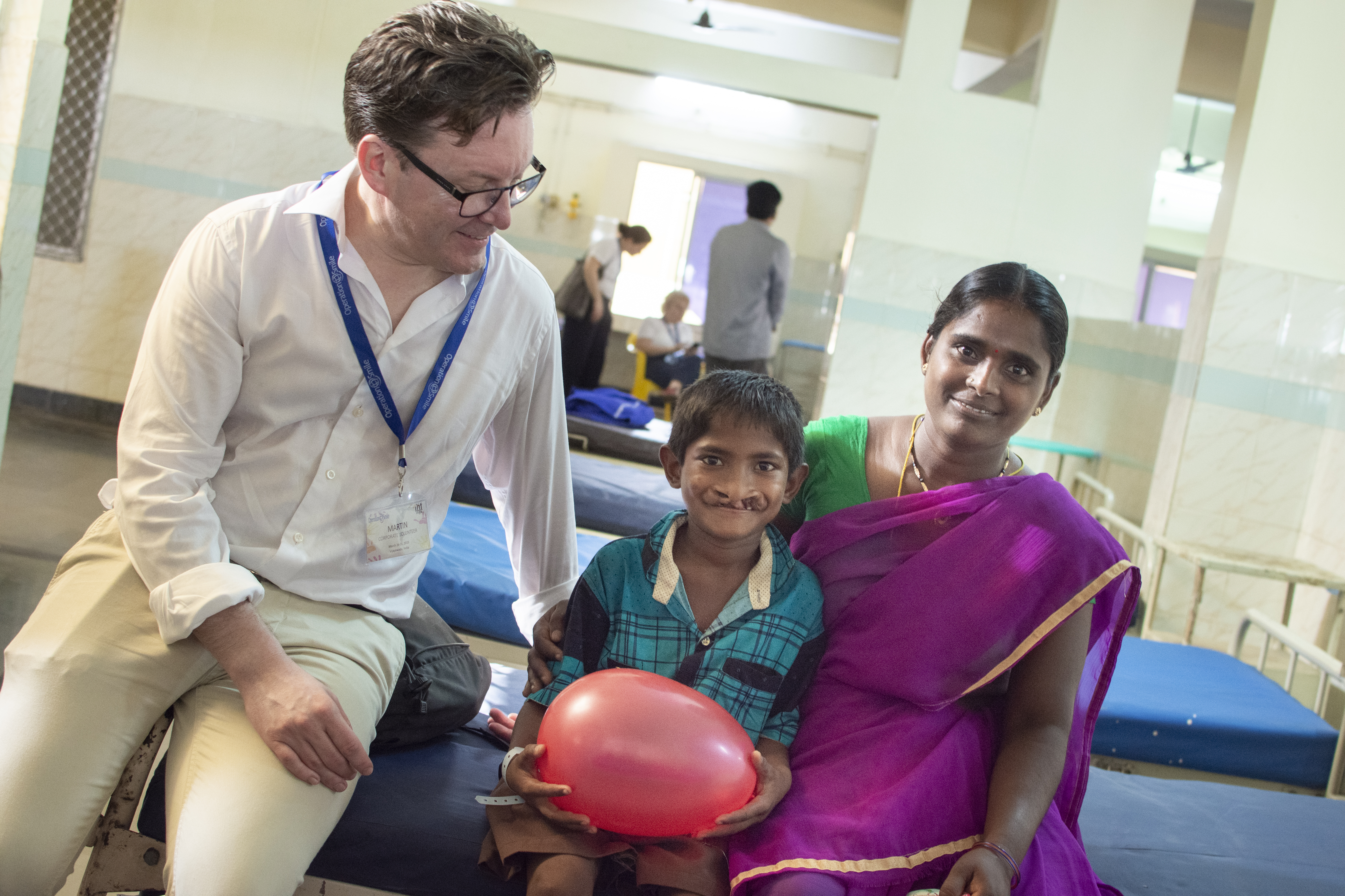 Martin Forsling med nyckelband runt halsen och besöksbricka sitter på en sjukhussäng bredvid en pojke med läpp-käk-gomspalt och hans mamma. Pojken håller i en ballong och han och hans mamma ler och tittar in i kameran.