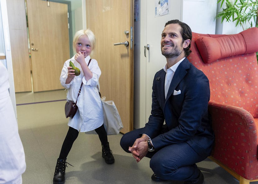Prins Carl Philip sitter på huk på golvet. Bredvid honom står en ljushårig flicka i femårsåldern som äter på ett päron. Båda tittar på en person som står utanför bild, till vänster.