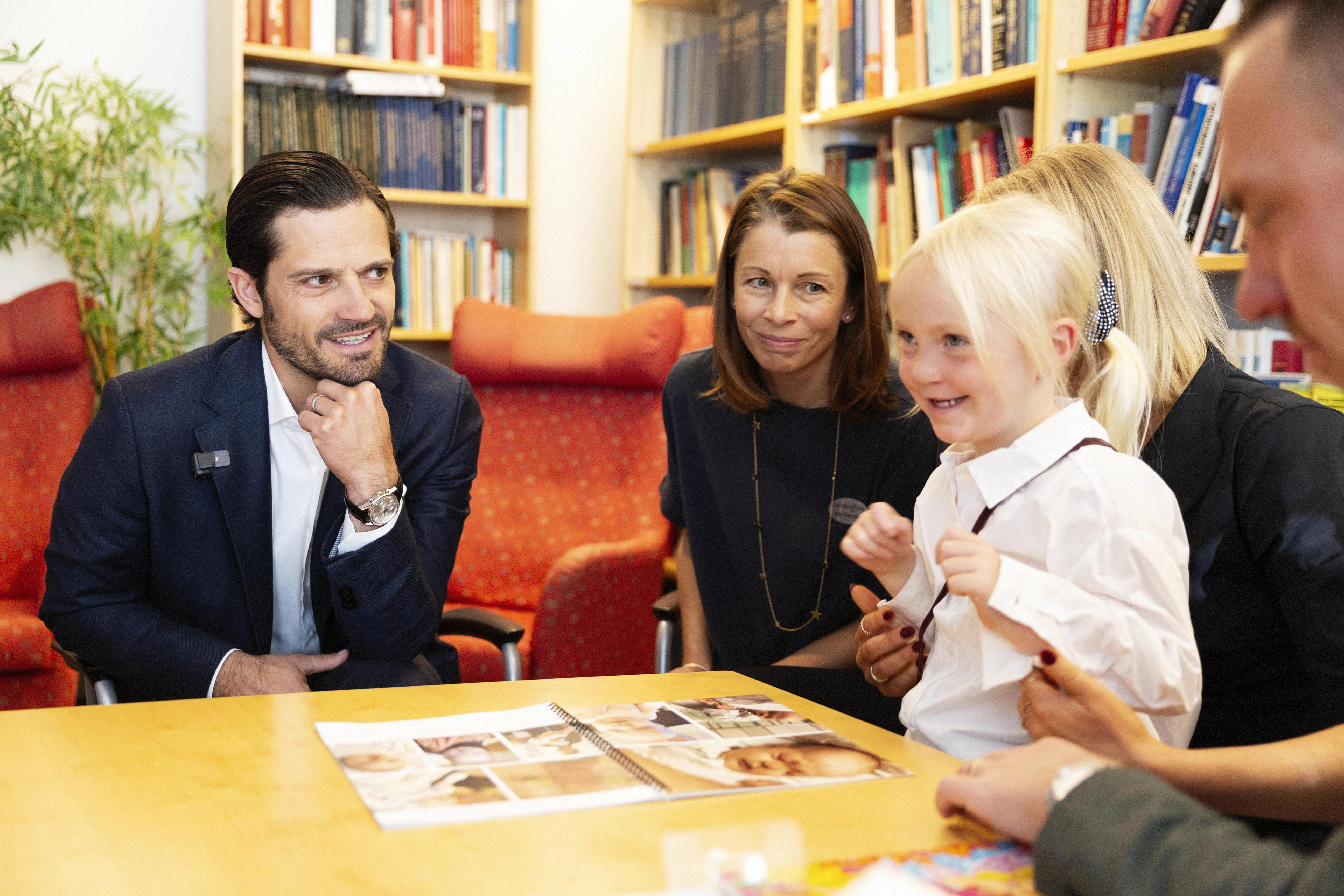 Prins Carl Philip och Malin Söderström, generalsekreterare, tittar på femåriga Emelinn som ser glad ut. Hon visar sitt fotoalbum med bilder från när hon föddes.
