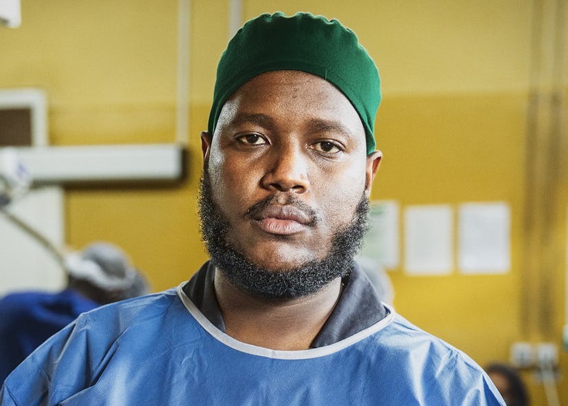 En man med grön kirurgmössa och blå arbetskläder står inomhus och tittar mot kameran med neutral min.